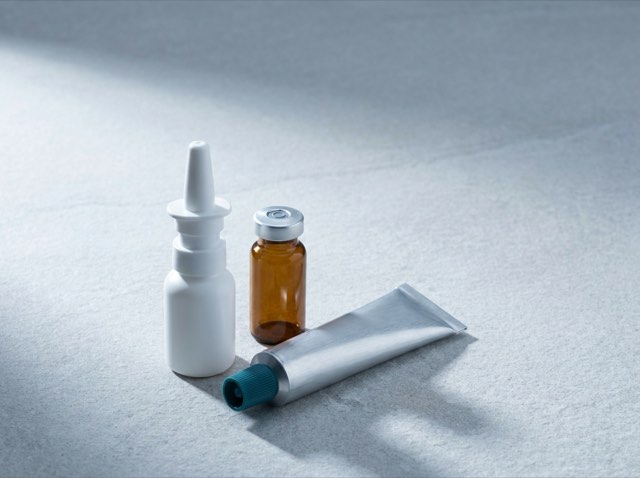 A nasal spray bottle, small amber vial and aluminium topical tube grouped on pale grey stone